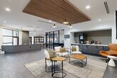 This single-floor amenity space offers a thoughtfully designed, multi-functional layout that balances collaborative and private uses. The open lounge area serves as the central hub, anchored by a linear fireplace and wall-mounted display screen. A striking slatted wood ceiling panel floats overhead, framed by recessed lighting and a multi-arm pendant fixture, adding warmth and architectural interest to the otherwise clean, white-walled interior. Large-format gray tile flooring flows throughout, lending a cohesive, contemporary feel.

Comfortable lounge seating — including white upholstered chairs, leather accent pieces, and woven area rugs — creates an inviting gathering area. Adjacent to the lounge, a built-in coffee and beverage bar with dark cabinetry provides a functional service station. A high-top communal table with bar seating occupies a separate area near a glass garage-style door that opens to an outdoor patio, promoting flexible indoor-outdoor use.

Two glass-walled conference rooms offer private meeting space with views to the outdoors. One room features floor-to-ceiling windows overlooking a landscaped exterior, filling the space with natural light. The overall design blends modern industrial elements with warm wood accents, creating a refined yet approachable amenity environment.