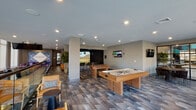 Located at 9 Tower Ln in New Haven, CT, this single-floor amenity space at Pierpont is designed to offer a sophisticated and engaging environment for recreation and socializing. The open-concept layout is anchored by floor-to-ceiling windows along the street-facing wall, flooding the interior with natural light and providing views of the surrounding New Haven streetscape. Durable, multi-toned plank flooring flows throughout, tying the various zones together cohesively. The game room area is a standout feature, equipped with an air hockey table, foosball table, and a golf simulator with a large projection screen — ideal for both casual fun and competitive play. A full-length shuffleboard table is positioned along the window wall, accompanied by pendant lighting that adds a warm, focused glow. Comfortable lounge seating, including leather swivel chairs and upholstered armchairs, is arranged throughout to encourage conversation and relaxation. A private meeting nook framed by floor-to-ceiling steel-and-glass partitions offers a quieter gathering space with a round table and plush seating. The kitchenette is finished with white cabinetry, quartz countertops, a matte black faucet, and a striking teal tile backsplash. Recessed lighting and a clean, neutral color palette complete the modern aesthetic throughout.
