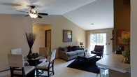 This single-floor residence showcases a thoughtfully designed open-concept layout that seamlessly connects the living and dining areas into one welcoming, cohesive space. The striking vaulted ceilings lend a sense of spaciousness and airiness, amplified further by a ceiling fan that adds both style and function overhead. The living area is anchored by a plush sofa and accent chair arrangement, centered around a dark wood coffee table and grounded by a bold area rug. A framed piece of wall art adds a touch of personality to the neutral-toned walls. Natural light pours in generously through a large sliding glass door, which offers views of the surrounding greenery and provides seamless access to an outdoor area. The dining space sits adjacent to the living room, featuring a round table with upholstered chairs and a decorative centerpiece, making it ideal for casual meals or intimate gatherings. Neutral carpeting throughout the shared space contributes to the warm, inviting ambiance. A hallway visible from the main area leads to additional rooms, hinting at a well-planned floor plan. Overall, the space balances comfort and modern aesthetics with clean lines and a harmonious color palette.