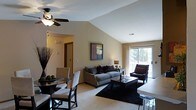 This single-floor residence showcases a thoughtfully designed open-concept layout that seamlessly connects the living and dining areas into one welcoming, cohesive space. The striking vaulted ceilings lend a sense of spaciousness and airiness, amplified further by a ceiling fan that adds both style and function overhead. The living area is anchored by a plush sofa and accent chair arrangement, centered around a dark wood coffee table and grounded by a bold area rug. A framed piece of wall art adds a touch of personality to the neutral-toned walls. Natural light pours in generously through a large sliding glass door, which offers views of the surrounding greenery and provides seamless access to an outdoor area. The dining space sits adjacent to the living room, featuring a round table with upholstered chairs and a decorative centerpiece, making it ideal for casual meals or intimate gatherings. Neutral carpeting throughout the shared space contributes to the warm, inviting ambiance. A hallway visible from the main area leads to additional rooms, hinting at a well-planned floor plan. Overall, the space balances comfort and modern aesthetics with clean lines and a harmonious color palette.