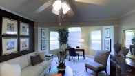 This single-story space centers around a versatile, open living area that seamlessly blends relaxation and functionality. The room's most distinctive architectural feature is its vaulted tray ceiling, which adds a sense of height and grandeur to the space. A ceiling fan with a decorative light fixture is mounted at the ceiling's peak, providing both airflow and ambient illumination.

Multiple windows, including a prominent arched window at the far end, allow generous amounts of natural light to pour in, brightening the warm, neutral-toned walls. The windowed alcove at the back of the room is thoughtfully arranged as a home office area, complete with a wooden desk, a navy blue accent chair, and a table lamp, making it an ideal workspace.

The main living area is furnished with a cream-colored sofa accented by patterned throw pillows, a round coffee table, and a cushioned armchair, all arranged atop a patterned area rug over carpeted flooring. A tall indoor fig tree and a potted floor plant add organic warmth and texture to the space. Framed artwork along one wall contributes to the polished, well-appointed ambiance of the room.