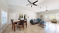 Located at 426 Mesa Ct in Little Rock, AR, this single-floor space offers an open, airy layout that seamlessly connects the living, dining, and kitchen areas. The interior is anchored by large-format travertine tile flooring throughout, complementing the neutral, light-toned walls that create a bright and welcoming atmosphere.

The expansive main living area features vaulted ceilings with crown molding and is equipped with multiple ceiling fans, enhancing both comfort and airflow. A wall-mounted television and ample seating make this space well-suited for gatherings. Natural light flows in generously through multiple windows and a glass-panel exterior door, offering views of the surrounding greenery.

The dining area is clearly defined, accommodating a solid wood pub-style table with upholstered chairs, and is bathed in natural light from a row of windows dressed with horizontal blinds. An open pass-through connects the dining space to the kitchen, which features cabinetry, a tile backsplash, and modern appliances visible from the main area.

Two interior doors are accessible from the main living space, indicated for restroom facilities, adding functional convenience. Decorative touches including a large wall clock, framed artwork, and accent lighting lend the space a polished, well-appointed character throughout.