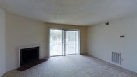 This single-floor space offers a comfortable and functional layout designed for everyday living. The main living area is a standout feature, characterized by plush wall-to-wall carpeting that adds warmth underfoot. A classic fireplace anchors one side of the room, providing a focal point and a cozy atmosphere during cooler months. Opposite the fireplace, a full-width sliding glass door fitted with vertical blinds floods the space with generous natural light and offers views of the surrounding outdoor greenery, creating a seamless connection between the interior and the outside environment. The neutral, light-toned walls and textured ceiling complement the open, airy feel of the room. The overall ambiance is clean, bright, and inviting, with a straightforward design that provides a versatile canvas for a variety of interior styles. The layout is well-suited for comfortable day-to-day living, with thoughtful details such as built-in ventilation and accessible wall outlets throughout.