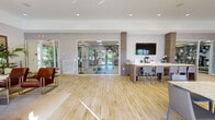 Located at 4400 NW 79th Ave in Doral, Florida, this single-story clubhouse and gym facility offers a well-appointed community gathering space designed with modern aesthetics and functional amenities. The interior is unified by warm light wood flooring, neutral wall tones, and wood-trimmed Roman shade windows that invite abundant natural light throughout.

The lounge area serves as a welcoming focal point, furnished with sleek black leather sofas, rich brown leather accent chairs, a glass-top coffee table, and a decorative area rug. Statement drum pendant lighting and lush tropical plants enhance the relaxed, upscale atmosphere. Adjacent to the lounge is a collaborative workspace featuring elegant marble-top tables with contemporary seating, a wall-mounted TV, and a built-in counter bar — ideal for residents to gather, work, or socialize.

Glass-paneled doors provide a seamless visual and physical connection to the fully equipped fitness center, which offers a comprehensive range of exercise equipment including treadmills, cable machines, weight benches, and a full dumbbell rack. Floor-to-ceiling mirrors and a bold kettlebell graphic mural energize the workout environment.

Additional doors open directly to the outdoor pool area, integrating indoor comfort with Florida's outdoor lifestyle. Overall, the space strikes a balance between community, comfort, and active living.