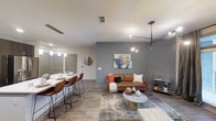 This single-floor residence showcases a thoughtfully designed open-concept layout that seamlessly connects the living and dining areas into one cohesive, functional space. The living area is anchored by a rich cognac leather sofa accented with bold throw pillows, set atop a layered area rug in muted tones. A striking multi-arm pendant light fixture serves as an artistic focal point overhead, adding an industrial-modern flair to the space. The accent wall, painted in a cool slate blue, provides visual depth and serves as a backdrop for decorative wall art, enhancing the contemporary aesthetic. Adjacent to the living area, a sleek bar-style dining counter with leather barstool seating offers a casual yet refined dining experience. Pendant lights hang above the counter, creating an intimate atmosphere for meals and entertaining. The flooring throughout features durable wood-look planks in a warm, neutral tone that ties the spaces together cohesively. Recessed lighting complements the pendant fixtures, ensuring the space is well-lit and versatile. The open flow, modern finishes, and carefully curated décor combine to create a stylish and welcoming single-floor living environment.