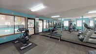 Located at 6000 65th Ave N in Brooklyn Park, Minnesota, this fitness center offers a well-equipped, single-floor workout space designed to support a variety of exercise routines. The room is immediately distinguished by its bold teal accent walls, which lend an energetic and motivating atmosphere throughout the space.

The cardio section features multiple treadmills, including Cybex-brand models, as well as stationary and recumbent bikes, providing options for users of varying fitness levels. For strength training, the facility includes a multi-station cable machine, a weight bench with free weights, and additional resistance equipment, giving members the tools for comprehensive full-body workouts.

Floor-to-ceiling mirrors line the walls, visually expanding the space and allowing users to monitor their form during exercise. The flooring consists of durable, multi-tone carpet tiles well-suited for a fitness environment. Overhead fluorescent lighting ensures the space is brightly and evenly lit throughout.

Natural light filters in through several windows along the perimeter walls, adding to the open and airy feel of the room. A glass door leads to an adjacent area, suggesting connectivity to additional amenities such as a pool. The textured ceiling and structural support columns contribute to the functional, practical design of this well-maintained fitness facility.