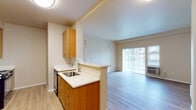 This single-floor residential unit offers a thoughtfully laid-out interior with a clean, functional design throughout. The heart of the unit is an open-concept kitchen and living area, where light hardwood-style flooring flows seamlessly underfoot. The galley-style kitchen is equipped with warm wood cabinetry, white countertops, a stainless steel sink, and black appliances including a refrigerator, stove, and dishwasher. A peninsula counter extends toward the living area, creating a natural divide while maintaining an open and connected feel. The living room is bright and airy, benefiting from a large sliding glass door that invites natural light and provides access to an outdoor area. A wall-mounted HVAC unit ensures year-round comfort. The bedroom features plush wall-to-wall carpeting, a window with exterior views, and a baseboard heater. The bathroom is outfitted with a wide vanity featuring a white countertop and wood cabinetry, a large mirror, and an accessible shower with grab bars. Completing the unit is a dedicated in-unit laundry area with a full-size washer and dryer, as well as additional storage space. Neutral warm tones throughout create a welcoming and cohesive ambiance.