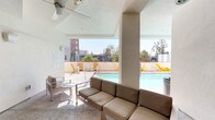 This outdoor pool deck is designed as a multifunctional amenity space that balances relaxation and social gathering. The centerpiece is a rectangular pool with clear turquoise water, bordered by a sun-exposed deck furnished with vibrant yellow lounge chairs and small side tables — a striking design choice that adds a bold, contemporary accent to the neutral palette of the surrounding architecture.

A covered cabana-style lounge area adjoins the pool, featuring polished terrazzo flooring, a ceiling fan overhead, and comfortable neutral-toned seating including a generously sized sofa and accent chairs. This shaded retreat opens directly onto the pool deck, creating a seamless indoor-outdoor flow. A wall-mounted television and a sleek fireplace clad in large-format dark grey tile serve as focal points within the covered section, enhancing the space's versatility for year-round use.

The deck is enclosed by a low perimeter wall that frames open views of the surrounding urban skyline, palm trees, and neighboring mid-rise buildings. Access to the pool area is controlled via a secured red metal gate. The overall ambiance is modern and resort-inspired, with a clean white stucco exterior and abundant natural light throughout.