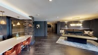 Located at 1350 Speer Blvd in Denver, Colorado, this single-floor amenity lounge is a masterfully designed social space that blends upscale aesthetics with leisure-focused functionality. The interior is anchored by a dramatic dark color palette — deep charcoal wall paneling, rich dark hardwood-style flooring, and bold accent walls — creating a moody yet inviting atmosphere throughout.

The games area serves as a central highlight, featuring a striking gold-framed billiards table set atop an ornate area rug, flanked by a built-in leather bench with teal accent pillows. Nearby, a dedicated ping-pong zone is illuminated by an elegant crystal chandelier with a brushed gold frame, set against a wall of decorative mirrors and paneled wainscoting.

The wet bar area is equally impressive, offering an extended white quartz countertop with seating for multiple guests, warm amber pendant lighting, open display shelving, and a built-in television — ideal for entertaining. Luxurious velvet bar chairs add a rich, tactile element to the space.

A lounge seating area near floor-to-ceiling windows allows natural light to filter in, offering views of the surrounding Denver neighborhood. The space is finished with curated artwork, sconce lighting, and layered rugs, making it a sophisticated retreat for residents and guests alike.