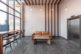 This contemporary single-floor lobby space makes an immediate impression with its dramatic double-height ceilings and sweeping floor-to-ceiling curtain glass walls that frame views of the surrounding streetscape, flooding the interior with natural light throughout the day. Dark-framed window systems lend an industrial refinement to the façade, while interior wood beam elements run across the ceiling and extend vertically down the walls, adding warmth and architectural rhythm to the otherwise crisp, neutral palette.

The main lounge area is anchored by a rich cognac leather sofa paired with a minimalist concrete coffee table, offering a relaxed yet polished seating vane. Complementing this is a high-top wooden bar table with industrial-style stools positioned directly against the glass wall, ideal for casual work or conversation with a street-facing view. Accent pendants with Edison-style bulbs hang at varying heights, contributing a warm, ambient glow.

A secondary seating nook features plush royal-blue velvet accent chairs grouped around a small side table, introducing a bold pop of color against the light gray walls and dark wood-look flooring. A stainless steel elevator with wood-trimmed framing serves as a functional focal point along one wall, accompanied by a pendant light overhead. The overall ambiance is modern, welcoming, and thoughtfully curated.