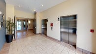 This commercial building lobby offers a polished and functional first impression, designed with accessibility and clarity in mind. The main entrance features full-height glass double doors framed in dark metal, flooding the space with natural light and offering unobstructed views of the surrounding greenery and outdoor environment. Light-toned ceramic tile flooring extends seamlessly throughout, lending a clean, cohesive aesthetic to the interior.

A bold slate-blue accent wall serves as a focal point near the entry, adorned with a large numeral "1" in brushed metal, a framed artwork piece, and essential building control panels. A tall potted bamboo plant adds a touch of organic warmth to the otherwise minimal palette. Wall-mounted sconces provide soft supplementary lighting alongside recessed ceiling fixtures.

The elevator lobby is centrally accessible, featuring two stainless steel elevator doors set against warm beige walls, accompanied by standard call panels and emergency signage. Floor-to-ceiling glazing adjacent to the elevators allows additional natural light to pour into the corridor, maintaining a bright and open atmosphere throughout.

The overall design balances practicality with a refined commercial aesthetic, creating a welcoming transition space that is clean, navigable, and well-maintained.