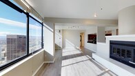 This single-floor unit at 3705 offers an open-concept layout filled with abundant natural light, courtesy of expansive floor-to-ceiling windows that frame striking panoramic city views. The living area features smooth light-toned hardwood flooring that runs throughout, complementing the clean white ceilings and neutral gray walls to create a bright, airy atmosphere. Sunlight streams across the space in dramatic patterns, highlighting the generous proportions of the open living and dining area. A kitchen sits at the rear of the space, distinguished by rich dark cabinetry and a breakfast bar with a contrasting white counter, offering a sleek visual anchor to the open floor plan. Track lighting overhead provides supplemental illumination in the kitchen area. A staircase with glass railing elements adds a contemporary architectural detail and contributes to the modern aesthetic. The overall ambiance is clean, minimalist, and urban, with the expansive city skyline visible through the large windows serving as a defining feature of the space.