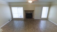 This two-story home offers a well-appointed layout designed for comfortable everyday living. The main living area serves as the centerpiece of the home, showcasing a classic fireplace framed by stone tile surround and a warm wood mantel — an inviting focal point for the room. Large windows on either side of the fireplace flood the space with natural light, creating a bright and airy atmosphere throughout. A ceiling fan with a light fixture is centrally positioned overhead, providing both comfort and illumination. The flooring throughout the living area consists of durable, neutral-toned ceramic tile laid in a grid pattern, offering both practicality and a clean aesthetic. Painted in soft, neutral tones, the walls complement the overall understated and functional design of the home. A doorway adjacent to the fireplace provides access to additional areas of the home, hinting at a well-connected floor plan. Spanning two floors, this home provides ample space for a variety of living arrangements, with the upper level offering additional rooms that expand the home's overall functionality. The combination of natural light, classic finishes, and a thoughtful layout makes this a comfortable and inviting space.