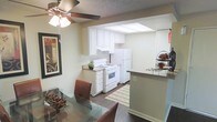 This single-story residence showcases a practical and well-organized open-concept layout, seamlessly connecting the dining area to the kitchen. The dining space is anchored by a glass-top table with warm brown chairs and a ceiling fan with integrated lighting overhead, adding both comfort and airflow to the area. A framed piece of wall art adds a decorative touch to the neutral-toned walls.

The kitchen is efficiently designed, featuring white cabinetry that extends to the upper walls, providing generous storage. White appliances, including a gas range with oven and a full-size refrigerator, are neatly arranged beneath a bright skylight-style overhead fixture that fills the space with ample illumination. A peninsula counter with a dark surface serves as a natural divider between the kitchen and dining areas, offering additional prep or serving space. Dark hardwood-style flooring runs throughout, adding warmth and visual continuity, while a patterned area rug in the kitchen zone adds texture. The overall ambiance is clean, neutral, and functional, making this a comfortable and livable home.