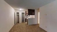 This single-floor unit offers a practical and efficiently designed layout that maximizes the use of its compact footprint. The living area features neutral-toned carpeting that extends throughout, creating a cohesive and understated aesthetic. A textured ceiling adds subtle visual character to the space.

The kitchen is positioned at the center of the unit and is defined by a white peninsula counter that serves as a natural divider between the kitchen and living area. Dark wood cabinetry overhead provides ample storage and contrasts with the white appliances, which include a refrigerator, gas range, and microwave. The kitchen floor transitions to a hard surface, distinguishing it from the carpeted living space.

Adjacent to the kitchen, a built-in open shelving unit offers additional storage and display space, adding functional versatility to the layout. A door nearby leads to a walk-in closet equipped with built-in shelving and a hanging rod, providing organized storage solutions. A hallway visible on the right side of the space leads to additional areas of the unit, including what appears to be a bathroom.