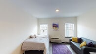 This studio unit at 933 N Walnut St in Bloomington, Indiana offers a thoughtfully designed single-floor layout that efficiently combines living, sleeping, and dining functions into one cohesive space. The main room is bright and open, featuring neutral walls, recessed lighting, and multiple windows fitted with blinds that invite natural light while maintaining privacy. A wall-mounted PTAC unit provides climate control, and the hard-surface flooring is complemented by a plush area rug that defines the living zone. The sleeping area accommodates a full-sized bed, while a grey sofa with accent pillows in yellow and purple provides a comfortable seating area nearby. A dedicated study nook includes a desk and chair, making the space practical for work or study. The kitchenette is equipped with dark espresso cabinetry, granite-style countertops, an under-counter refrigerator, and a countertop oven — offering convenient self-catering capabilities. The private bathroom features matching dark cabinetry with a granite-top vanity, an undermount sink, a large mirror, and a shower. The overall aesthetic is clean and modern, with a consistent dark-and-light contrast throughout, making this a functional and comfortable space ideally suited for individual living near Indiana University.