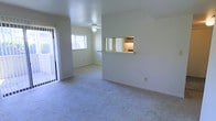 This single-story space features an open and functional layout designed to maximize usability. The living area is finished with wall-to-wall carpet flooring in a neutral tone, creating a clean and cohesive look throughout. A ceiling fan is centrally positioned overhead, providing both light and air circulation to keep the space comfortable year-round.

One of the notable architectural details is a pass-through window that connects the kitchen to the main living area. Framed with a light wood ledge, this opening enhances the sense of connectivity between spaces while allowing natural light from the kitchen to filter through. The kitchen itself is partially visible through this opening, suggesting a compact and efficiently arranged cooking space.

Natural light plays an important role in the ambiance of this unit. A large window in the adjoining room draws in generous daylight, while a sliding glass door on the opposite wall offers direct access to an outdoor area, further brightening the interior. The walls are finished in a crisp, neutral white, complementing the light tones of the carpet and contributing to an overall bright and airy atmosphere throughout the space.