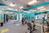 Located at 151 Grande View Dr in Biloxi, Mississippi, this single-floor fitness center delivers a well-equipped and energizing workout environment with a distinctly coastal aesthetic. The space is defined by bold teal accent walls that create a vibrant, motivating atmosphere, complemented by a dramatic floor-to-ceiling ocean wave mural that serves as a striking focal point and reinforces the Gulf Coast character of the facility.

The layout is open and generously proportioned, allowing for comfortable movement between equipment zones. Full-length mirrors line key sections of the walls, visually expanding the space and providing users with form-checking capability throughout their workouts. Overhead, vaulted ceiling sections are fitted with ceiling fans and recessed fluorescent lighting, ensuring the room remains well-lit and well-ventilated.

The cardio zone is outfitted with treadmills, upright and recumbent Matrix bikes, and a spinning bike, positioned near large windows that provide natural light and views of the surrounding palm-lined outdoor area. The strength training area includes a cable machine, weight bench, free weights, and additional resistance equipment. A wall-mounted television, a water fountain, and designated floor markers for stretching or mat work round out the amenities, making this a fully functional fitness facility.