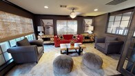 Located at 1500 Plantation Oaks Dr in Trophy Club, Texas, this well-appointed single-story community space offers a cohesive and sophisticated interior designed for both relaxed socializing and organized gatherings. The layout flows seamlessly between distinct functional areas, each thoughtfully finished with warm, contemporary décor.

The lounge area anchors the space with rich, dark-toned walls, recessed lighting, and large woven Roman shades that filter natural light from generous windows. A bold red tufted sofa serves as the focal point, complemented by classic gray armchairs, rounded upholstered ottomans, a rustic wood-and-metal coffee table, and matching credenzas flanked by framed artwork.

An elegant set of dark wood French doors with glass panes connects the lounge to the main gathering hall, creating a grand transition between spaces. The kitchen and bar area features dark hardwood flooring, a long granite-topped island with red leather bar stools, full appliances, and striking geometric wood art panels on the walls. A chandelier and arched window brighten the entry corridor beyond.

The dining area accommodates a large wooden table with mixed seating, surrounded by woven shades and decorative wall art. Tile flooring and recessed lighting create a refined yet comfortable atmosphere throughout. This versatile space is well-suited for community events, private functions, and everyday resident use.