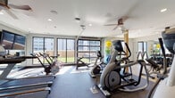 Located at 222 Sycamore St in Evansville, Indiana, this fitness center is a comprehensively outfitted single-floor facility designed to support a full range of workout routines. The space is divided into distinct functional zones, each thoughtfully arranged to maximize usability and flow.

The cardio area is positioned along a wall of large, floor-to-ceiling windows and glass garage-style doors that flood the room with natural light and provide views of the surrounding urban environment. This section houses multiple Matrix treadmills, stationary cycling bikes, elliptical trainers, and a stair climber, all arranged to take advantage of the bright, airy atmosphere. Ceiling fans overhead help maintain comfortable airflow throughout the space.

The strength training zone features full mirror-lined walls that visually expand the room and aid in form correction. A well-stocked dumbbell rack with colorful kettlebells, tiered weight racks, a weight bench, and selectorized strength machines are neatly organized in this area. Natural wood accent columns add a warm architectural contrast to the otherwise modern, neutral-toned interior.

A secondary area near the entrance provides built-in shelving and additional equipment storage, alongside a water fountain and direct access via a wood-paneled door. Rubber and speckled flooring throughout ensures safety and durability, completing a practical and polished fitness environment.
