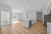 This contemporary single-floor residence is designed with a clean, modern aesthetic and a bright, open layout. The heart of the space is a spacious open-concept kitchen and living area, unified by light-toned hardwood flooring that extends throughout. The kitchen is well-appointed with white shaker-style cabinetry featuring matte black hardware, a large center island with a white quartz countertop and undermount sink, and a full suite of stainless steel appliances including a refrigerator, range, over-the-range microwave, and dishwasher. A trio of globe pendant lights hang above the island, adding a modern decorative touch. The adjacent living area is generously sized, bathed in natural light, and opens directly to an outdoor balcony through sliding glass doors, offering scenic views of the surrounding landscape. The bedrooms are carpeted for comfort and each features large windows that allow ample daylight and outdoor views. The full bathroom is finished in a clean, all-white palette with a combined tub and shower, a vanity with sink, and hardwood-style flooring. Throughout the unit, neutral warm-white walls and consistent finishes create a cohesive, move-in-ready atmosphere.