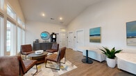 Located at 2052 Kali Oka Rd in Saraland, Alabama, this single-floor living space offers a thoughtfully designed communal lounge environment. The room features vaulted ceilings that amplify the sense of openness, while warm-toned luxury vinyl plank flooring runs throughout, grounding the space with a clean, contemporary aesthetic.

A central seating arrangement anchors the room, consisting of rich brown leather chairs grouped around a low round coffee table atop a geometric patterned area rug in muted yellows, grays, and blues. A white curved sofa and a sleek pedestal table with a marble-look top provide additional seating and social areas along one wall.

The accent wall in soft blue-gray adds a calming contrast to the surrounding white walls, which are adorned with coastal-themed artwork. A wall-mounted flat-screen television serves as a focal point on this accent wall. Recessed lighting and natural light from a large multi-pane window wall and glass door ensemble illuminate the space, offering views into an adjacent fitness area visible through the glazing.

A built-in gray cabinetry station with a granite countertop, circular mirror, and beverage machine serves as a refreshment area. Interior glass-panel doors lead to additional rooms. Potted greenery is positioned throughout, enhancing the fresh, welcoming ambiance of this well-curated space.