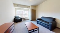 This single-floor unit offers a practical and well-organized layout designed for comfortable everyday living. The open-concept kitchen and living area form the heart of the space, with the kitchen featuring warm wood cabinetry, dark granite-style countertops, and a full suite of black appliances including a range, microwave, dishwasher, and refrigerator. A peninsula with bar-height seating provides a casual dining option and visually connects the kitchen to the adjoining living area.

The living room is carpeted and bright, receiving natural light through a broad window that offers views of the surrounding exterior. The space comfortably accommodates a sofa, armchair, and coffee table arrangement, making it well-suited for both relaxing and socializing.

The bedrooms are carpeted and modestly sized, each offering enough room for a bed and a desk-and-chair setup. Windows in the bedrooms allow for natural light and exterior views. The bathroom includes a single-sink vanity with wood cabinetry, a full tub-shower combination, and light-toned flooring throughout. The overall aesthetic is clean, neutral, and functional, with a layout that makes efficient use of the available space.