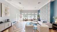 Located in Houston, TX (77089), this thoughtfully designed single-floor space comprises a generously sized living room and a connecting hallway. The living room immediately draws the eye with its striking teal accent walls, crisp white crown molding, and a dramatic floor-to-ceiling decorative wallpaper panel that serves as a bold focal point. Flanking the panel, elegant wall sconces cast a warm, inviting glow throughout the room. The space is anchored by a stunning herringbone-patterned hardwood floor that extends seamlessly into the hallway, lending a sense of refined continuity. Natural light filters in through windows, brightening the open layout and complementing the cool, coastal-inspired color palette of blues and grays. The living area is further appointed with console tables, round mirrors, and curated artwork, while a mounted flat-screen television and credenza add functional versatility. The hallway connects to additional areas via wide doorways, enhanced by a chandelier overhead and artwork along the walls. Together, the rooms create a cohesive, welcoming atmosphere that balances modern elegance with warm, livable comfort.