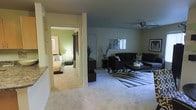 This single-floor residence presents a well-organized layout connecting a spacious living area to a private bedroom. The living room is furnished with a dark sectional sofa and coordinating accent chairs, arranged around a sleek coffee table atop a bold black-and-white striped area rug, lending a contemporary feel to the space. A ceiling fan with an integrated light fixture is centrally mounted overhead, providing both airflow and ambient lighting. Decorative wall art and a framed piece add visual interest to the neutral-toned walls. A tall potted plant introduces a natural element, complementing the room's modern aesthetic. The flooring throughout is plush carpeting in a neutral tone, contributing to a warm and comfortable atmosphere. A glimpse into the adjoining bedroom reveals green accent walls, soft bedding, and a bedside lamp, creating a relaxed, intimate retreat. A dark console table near the entryway offers functional storage, and a white interior door provides access to additional areas of the residence. The overall ambiance is contemporary and inviting, with a cohesive color palette and thoughtful furnishing choices throughout.