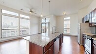 This well-appointed single-floor urban unit showcases a thoughtfully designed open layout with soaring ceilings that amplify the sense of space throughout the main living areas. The kitchen serves as a central hub, featuring a large granite-topped island with an undermount sink and pendant lighting, rich dark-wood cabinetry extending to the ceiling, subway tile backsplash, and a full suite of stainless steel appliances including a refrigerator, range, and built-in microwave. Hardwood flooring flows seamlessly through the kitchen and living areas, adding warmth and continuity.

The living area is defined by dramatic floor-to-ceiling windows and transom windows above, flooding the space with abundant natural light while framing panoramic views of the Atlanta skyline. A ceiling fan adds comfort, and a sliding glass door provides direct access to a private balcony overlooking a tree-lined urban landscape with striking city views.

The bedrooms are carpeted for comfort and feature large windows that invite natural light. A spacious walk-in closet with wire shelving offers generous storage. The bathroom is finished with tile flooring, a tiled soaking tub surround, and dark-wood vanity cabinetry consistent with the unit's overall design palette.