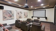 This private screening room is thoughtfully designed to deliver a premium cinematic experience. At the heart of the space is a large projection screen mounted on the front wall, paired with a ceiling-mounted projector that ensures a crisp, high-quality picture. The seating arrangement features rows of generously sized, plush armchairs upholstered in rich charcoal fabric, each accented with red throw pillows for a touch of elegance and comfort. The chairs are arranged to provide unobstructed sightlines to the screen from every seat. The room is finished with warm, neutral-toned walls adorned with two large abstract art pieces, adding a sophisticated, gallery-like quality to the space. A tray ceiling with recessed lighting and dark wood crown molding frames the room, creating an intimate and upscale atmosphere. The patterned area rug anchors the seating area and complements the overall color palette. Together, the thoughtful layout, high-end furnishings, and dedicated audiovisual equipment make this screening room an ideal venue for private movie viewings and entertainment gatherings.