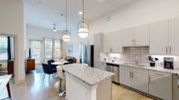 This contemporary single-floor residence showcases a thoughtfully designed open-concept layout that seamlessly connects the kitchen, dining, and living areas. Soaring ceilings and polished concrete floors run throughout, lending the space an airy, loft-like character. The kitchen is a standout feature, equipped with stainless steel appliances, gray flat-front cabinetry with gold hardware, a white subway tile backsplash, and a granite-topped island with bar seating illuminated by stylish brass pendant lights. A dedicated dining area with a round table flows naturally into the living room, where a ceiling fan, large windows, and a glass door bring in generous natural light and views of the surrounding greenery. The entryway includes a built-in console area and a circular gold-framed mirror, offering a welcoming first impression. Double doors near the entry provide additional closet or utility storage. The bedroom is well-proportioned, featuring a large window with views, high ceilings, and polished concrete flooring. The bathroom includes a tub-shower combination with marble-style tile surround, granite vanity countertop, and direct access to a walk-in closet outfitted with multi-level shelving and hanging rods. The overall aesthetic is clean, modern, and cohesive throughout.