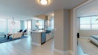 This well-appointed single-floor residence showcases a thoughtfully designed open-concept layout bathed in natural light. The kitchen serves as a central highlight, featuring white shaker cabinetry, quartz countertops, a farmhouse-style sink, subway tile backsplash, and a full suite of stainless steel appliances including a dishwasher, range, microwave, and French door refrigerator. A peninsula with bar seating provides additional prep space and casual dining. Adjacent to the kitchen, a dedicated dining nook accommodates a round table beneath a statement pendant light, with large windows framing expansive outdoor views.

The living area is open and airy, anchored by floor-to-ceiling sliding glass doors that connect seamlessly to a balcony offering panoramic views. Light-toned tile flooring extends throughout, enhancing the sense of space. Two bathrooms feature glass-enclosed walk-in showers with large-format tile surrounds, white vanities with quartz countertops, and well-lit mirrors. The bedroom is calm and spacious, finished with soft neutral tones and elevated by a striking star-shaped chandelier. Overall, the residence presents a cohesive, contemporary aesthetic with abundant natural light and refined detailing.