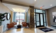 This single-floor space presents a polished and contemporary lobby designed to make a strong first impression. The entrance area is anchored by a striking decorative column clad in an intricate laser-cut metallic screen pattern, serving as a bold architectural focal point that draws the eye immediately upon entry. Large floor-to-ceiling windows flood the interior with abundant natural light, creating an open and airy ambiance throughout the space.

A comfortable lounge seating area is thoughtfully arranged near the windows, featuring rich cognac leather armchairs paired with a sleek, dark round coffee table and accented by a patterned area rug in deep blue tones. An elegant multi-arm chandelier with exposed bulbs adds a modern yet refined lighting element above the seating arrangement.

The flooring is composed of large-format light-toned tiles that enhance the sense of openness, while a recessed tray ceiling detail above the lounge area adds architectural depth and dimension. The main entrance features a set of full-glass doors framed in dark metal, reinforcing the building's modern aesthetic. Overall, the space balances sophistication with functionality, offering a bright, inviting environment suited for a professional or commercial setting.