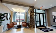This single-floor space presents a polished and contemporary lobby designed to make a strong first impression. The entrance area is anchored by a striking decorative column clad in an intricate laser-cut metallic screen pattern, serving as a bold architectural focal point that draws the eye immediately upon entry. Large floor-to-ceiling windows flood the interior with abundant natural light, creating an open and airy ambiance throughout the space.

A comfortable lounge seating area is thoughtfully arranged near the windows, featuring rich cognac leather armchairs paired with a sleek, dark round coffee table and accented by a patterned area rug in deep blue tones. An elegant multi-arm chandelier with exposed bulbs adds a modern yet refined lighting element above the seating arrangement.

The flooring is composed of large-format light-toned tiles that enhance the sense of openness, while a recessed tray ceiling detail above the lounge area adds architectural depth and dimension. The main entrance features a set of full-glass doors framed in dark metal, reinforcing the building's modern aesthetic. Overall, the space balances sophistication with functionality, offering a bright, inviting environment suited for a professional or commercial setting.