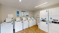 Located at 550 Crossway Ave in Murfreesboro, Tennessee, this shared laundry facility offers a clean, well-organized, and efficiently designed space for residents. The room is laid out to maximize usability, featuring a row of top-loading washing machines along one wall and three stacked dryer units along the adjacent wall, providing ample capacity for multiple users. All machines are equipped with modern card-based payment systems, offering a convenient and cashless laundry experience. The space is finished with warm wood-look luxury vinyl flooring and neutral-toned walls, lending a tidy and welcoming atmosphere. Overhead linear fluorescent lighting ensures the room is well-lit throughout. A wall-mounted air conditioning unit provides climate control for year-round comfort. Safety is addressed with a visible fire extinguisher and an electrical panel mounted on the wall. Informational laundry tip posters and a notice board are displayed, offering helpful guidance to users. The overall layout is practical and uncluttered, making the laundry process straightforward and accessible for all residents.