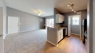 This single-floor, one-bedroom, one-bathroom residence offers a thoughtfully designed open-concept layout finished in neutral gray tones throughout. The living area features plush carpeting and a ceiling fan, with sliding glass doors leading to a private balcony that brings in natural light and outdoor views. Ceiling fan fixtures are present in both the living area and bedroom, contributing to comfortable airflow throughout.

The kitchen is defined by a peninsula with granite countertops, white cabinetry, and track lighting overhead. Black appliances, including a gas range and stainless steel refrigerator, complement the space, while hardwood-style flooring grounds the cooking and dining area.

The bedroom is generously proportioned with soft carpeting and features a sliding-door closet outfitted with built-in shelving and hanging rods, providing ample organized storage.

The bathroom includes a full soaking tub with tile surround, a white vanity with granite countertop, overhead cabinetry above the toilet for additional storage, and hardwood-style flooring.

A dedicated laundry room with a full-size washer and dryer, hardwood-style flooring, and a window rounds out this well-appointed residence.