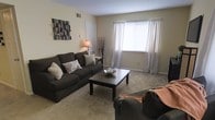 This single-floor residence offers a comfortable and inviting atmosphere with a thoughtful layout designed for everyday living. The living room serves as the heart of the space, showcasing plush wall-to-wall carpeting in a warm neutral tone that adds softness underfoot. Large windows dressed with sheer white curtains allow abundant natural light to fill the room, creating an airy and open feel throughout the day. The walls are finished in a soft, warm beige, contributing to the overall cozy ambiance. A decorative wall-mounted art piece adds a touch of character and visual interest to the space. The room comfortably accommodates generous seating arrangements, complemented by a classic dark-finished coffee table that anchors the sitting area. Subtle decorative accents, including a floor lamp and tasteful tabletop décor, add warmth and personality without overwhelming the space. A ceiling vent ensures efficient climate control throughout. The overall design aesthetic is clean, understated, and functional, making the space well-suited for relaxed daily living and casual entertaining alike.