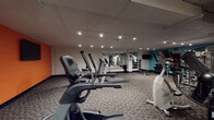This fitness center is a comprehensively outfitted single-floor exercise space designed to support a well-rounded workout routine. The room is finished with durable patterned carpet flooring and illuminated by recessed and track lighting mounted on the ceiling, providing consistent, functional brightness throughout the space.

The cardio section is well-stocked, featuring multiple treadmills, elliptical trainers, and stationary bikes from brands such as True Fitness, offering users a variety of aerobic exercise options. A dedicated strength training area includes a cable and weight machine, an adjustable workout bench, and a tiered dumbbell rack holding a full range of free weights. Exercise balls in yellow and red are also available, supporting flexibility and core training.

Large wall-mounted mirrors span one side of the room, visually expanding the space and allowing users to monitor their form during workouts. Wall-mounted flat-screen televisions are positioned throughout for entertainment. Bold teal and orange accent walls add a vibrant, energetic character to the otherwise neutral interior. The layout is organized to allow comfortable movement between equipment zones, making efficient use of the available floor space.