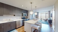 Located at Reston Station Boulevard in Reston, Virginia, this single-floor apartment offers a thoughtfully designed open-concept layout with contemporary finishes and abundant natural light. The kitchen serves as a central feature of the living space, showcasing dark wood-grain cabinetry that extends to the ceiling, a subway tile backsplash, quartz countertops, and a full suite of stainless steel appliances including a gas range, dishwasher, built-in microwave, and refrigerator. A white kitchen island with seating provides additional prep space and casual dining, while two pendant lantern-style light fixtures add a refined touch overhead. The kitchen flows seamlessly into the living area, which features warm hardwood flooring, floor-to-ceiling windows that draw in generous natural light, and views of the surrounding area. The bedrooms are carpeted and similarly well-lit, each featuring large windows with blinds and sheer drapery. Two bathrooms serve the unit; one features bold teal accent walls, a wood-toned vanity, and a well-lit mirror, while the second is finished in neutral tones with tile flooring and a tub-shower combination. The overall design aesthetic is modern and cohesive throughout.