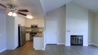 This single-story space showcases an open-concept layout that seamlessly connects the kitchen and living area. A defining architectural feature is the vaulted ceiling, which rises dramatically over the living space, creating a sense of openness and volume. A ceiling fan is positioned over the kitchen area for added comfort.

The kitchen is well-appointed with stainless steel appliances, including a refrigerator, gas range, and over-the-range microwave. White cabinetry lines the walls, providing ample storage, while dark countertops offer a contrasting visual element. A peninsula-style counter extends into the space, creating a natural boundary between the kitchen and living areas while offering additional prep or seating space.

The living area features a fireplace with a classic white mantel, serving as a focal point on one wall. Hardwood-style flooring runs throughout the open area, contributing warmth and continuity. A white paneled door provides access to what appears to be an exterior entry point. The neutral color palette across walls, trim, and cabinetry gives the space a clean, versatile aesthetic throughout.