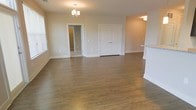 This single-floor residence offers a clean, modern aesthetic defined by warm hardwood-style flooring that flows seamlessly throughout the open living area. The space is bathed in natural light, courtesy of well-placed windows that illuminate the neutral, soft beige walls and crisp white trim. A elegant chandelier serves as a focal point overhead, adding a refined touch to the main living area.

The open layout creates a generous sense of space, allowing easy movement between rooms. A paneled white door leads to an additional room, while a separate doorway opens to what appears to be a hallway or secondary space, suggesting a thoughtful and functional floor plan. The raised counter or bar area visible to the side hints at a kitchen or dining transition zone, further emphasizing the home's versatile, connected design.

The overall ambiance is light, airy, and move-in ready, with a neutral palette that provides an ideal canvas for personalization. The combination of consistent flooring, subtle architectural details, and well-proportioned rooms gives this single-story home a welcoming and cohesive character throughout.