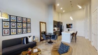 This single-floor residence is designed with an open-concept layout that integrates the living, dining, and kitchen spaces into one cohesive and functional area. The most striking architectural feature is the dramatically high ceilings, which lend an expansive, airy quality to the interior and amplify the sense of space throughout.

The living area is anchored by a sleek sofa and complementary seating, centered around a unique live-edge wood coffee table that adds a natural, organic contrast to the otherwise contemporary design. Recessed lighting and light wood-tone flooring run throughout, creating a warm yet modern ambiance.

The dining space sits at the heart of the open floor plan, furnished with a round table and plush velvet chairs, providing an intimate setting within the larger open area. A full-length mirror adds depth and dimension to this zone.

The kitchen is outfitted with dark cabinetry, stainless steel appliances, and a bright white peninsula countertop that doubles as a breakfast bar, complete with modern bar seating. Elegant pendant lights hang above the peninsula, providing focused task lighting while serving as a stylish design element.

Overall, the space exhibits a sophisticated urban aesthetic, combining bold design choices with practical functionality in a bright, well-lit environment.