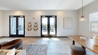Located at 1827 N Meridian St in Indianapolis, Indiana, this thoughtfully designed community lounge space combines modern aesthetics with warm, natural materials across a single open floor. The layout seamlessly integrates a living room, dining area, and kitchen into one cohesive, flowing environment.

A defining architectural feature throughout the space is the striking reclaimed wood accent wall, which adds texture and warmth against the crisp white walls and light-toned luxury vinyl plank flooring. The kitchen is outfitted with white shaker cabinetry, a stone tile backsplash, sleek light countertops, and stainless steel appliances including a dishwasher, microwave, and compact refrigerator — all framed by the same wood paneling for a unified look.

The dining area is anchored by a solid wood table beneath a trio of industrial-style pendant lights, with windows offering views of the surrounding Indianapolis cityscape. The living room features comfortable seating, a wall-mounted television, and an area rug that grounds the space.

Black-framed French doors open to an outdoor terrace, flooding the interior with natural light and providing panoramic views of the Indianapolis skyline. Decorative wall art and carefully curated furnishings complete the inviting, contemporary ambiance of this well-appointed communal space.