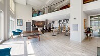 Located at 3011 Hidden Forest Ct in Marietta, Georgia, this single-floor community clubhouse offers a generously sized, thoughtfully designed gathering space that blends modern aesthetics with functional amenities. The entrance welcomes visitors with floor-to-ceiling glass walls and doors that flood the interior with natural light while framing views of the outdoor courtyard and pool area beyond.

The soaring double-height ceilings create an open, airy atmosphere throughout the living room, which serves as the social heart of the space. Light wood-look tile flooring runs seamlessly across the main area, complementing the bright white walls and contemporary furnishings. Stylish teal accent chairs and a blue sectional sofa are arranged around a marble-top coffee table, providing inviting seating nodes for casual conversation or relaxation.

A ping-pong table anchors one section of the room, adding a recreational element to the space. Nearby, a wall-mounted flat-screen TV is set into a recessed niche flanked by additional lounge seating. Toward the back, a dark-tiled kitchenette with a bar counter and pendant lighting serves as a refreshment hub, adjacent to a co-working café area filled with high-top tables and bistro chairs beneath bold typographic wall art. An upper-level mezzanine with a metal railing overlooks the main floor, adding architectural depth and visual interest to this dynamic, community-focused space.