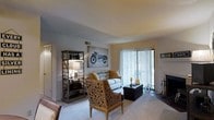 This single-floor space offers a comfortable and thoughtfully designed living environment. The living area serves as the focal point of the home, featuring plush wall-to-wall carpeting that adds warmth underfoot. Large sliding glass doors dressed with vertical blinds allow generous natural light to fill the room while providing a seamless connection to the outdoors. The space is enhanced by smooth, neutral-toned walls and ceilings that create a bright and airy atmosphere throughout. The living area is well-proportioned, offering ample room for both relaxed seating and functional living. An open-concept layout promotes a natural flow between spaces. Distinctive design elements include an ornate, laser-cut accent chair that serves as a bold visual centerpiece, complemented by a classic sofa with warm accent pillows. An industrial-style open shelving unit adds both storage and character to the room. A fireplace with traditional ironwork accessories anchors one corner of the space, adding a cozy and inviting element. Overall, the space strikes a balance between comfort and character, making it an inviting and functional home.