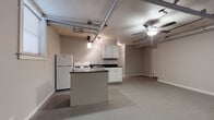 This single-floor unit showcases an industrial loft aesthetic, highlighted by exposed overhead pipes and ductwork running across the raw ceiling — a defining architectural feature that gives the space a distinctive urban character. Ceiling fans are positioned throughout to ensure comfortable airflow across the open layout.

The kitchen is well-equipped with white cabinetry, dark countertops, a gas range, a full-size refrigerator, and a stainless steel sink. A central island with a beadboard panel provides additional counter space and storage, serving as a natural divider between the kitchen and the generous open living area. The flooring transitions between tile and carpet, grounding the different functional zones of the space.

The living area is notably spacious, offering flexibility for various furniture arrangements. Natural light filters in through multiple windows, lending warmth to the otherwise industrial setting. Pendant lighting adds a soft, ambient glow to complement the overhead ceiling fan light fixture.

A full bathroom includes a combination tub and shower with a curtain enclosure, a toilet, and tile flooring, providing all essential amenities. Additional cabinetry and storage areas are distributed throughout the unit. Overall, this space balances raw, industrial design elements with practical, comfortable living features.