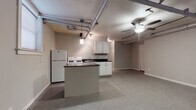 This single-floor unit showcases an industrial loft aesthetic, highlighted by exposed overhead pipes and ductwork running across the raw ceiling — a defining architectural feature that gives the space a distinctive urban character. Ceiling fans are positioned throughout to ensure comfortable airflow across the open layout.

The kitchen is well-equipped with white cabinetry, dark countertops, a gas range, a full-size refrigerator, and a stainless steel sink. A central island with a beadboard panel provides additional counter space and storage, serving as a natural divider between the kitchen and the generous open living area. The flooring transitions between tile and carpet, grounding the different functional zones of the space.

The living area is notably spacious, offering flexibility for various furniture arrangements. Natural light filters in through multiple windows, lending warmth to the otherwise industrial setting. Pendant lighting adds a soft, ambient glow to complement the overhead ceiling fan light fixture.

A full bathroom includes a combination tub and shower with a curtain enclosure, a toilet, and tile flooring, providing all essential amenities. Additional cabinetry and storage areas are distributed throughout the unit. Overall, this space balances raw, industrial design elements with practical, comfortable living features.