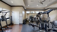 Located at 4101 W Huntington Dr in Rogers, Arkansas, this well-appointed fitness center offers a thoughtfully designed single-floor workout space with a generous footprint. The room features an elegant tray ceiling accented by dark crown molding, recessed lighting, and a ceiling fan, creating a polished yet functional atmosphere. Large windows along multiple walls flood the space with natural light while providing views of the surrounding exterior, enhancing the overall workout environment. Full-length mirrors are strategically placed to visually expand the space and assist with exercise form. The flooring transitions between durable rubber sport flooring in the main workout areas and hardwood-style flooring in a dedicated functional training zone. Equipment is comprehensive and well-organized, including multiple treadmills, elliptical machines, and a stationary bike for cardiovascular training, alongside a multi-station cable machine, a power tower, free weight racks with dumbbells, and benches for strength training. Additional functional fitness tools such as stability balls and medicine balls round out the offerings. A water cooler is conveniently available on-site. The facility is accessible via both an interior door and an exterior entrance, with posted fitness center rules ensuring a well-managed environment for all users.