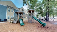 Located at 8439 Dorchester Rd in North Charleston, South Carolina, this outdoor recreational space features a well-appointed playground designed for active play and outdoor enjoyment. The centerpiece of the area is a multi-tower wooden play structure, consisting of two gazebo-style towers connected by a central climbing wall panel outfitted with colorful handholds. Two green slides extend from the towers, offering children multiple play options. A set of wooden stairs with railings provides accessible entry to one of the towers, while the climbing wall adds an adventurous alternative route. The entire play area is surfaced with a generous layer of wood chip mulch, providing a soft, safety-conscious ground cover beneath the equipment. Mature trees surround the space, offering natural shade and a lush, green canopy overhead, contributing to a comfortable outdoor environment. In the background, multi-story residential buildings with siding in complementary shades of blue, yellow, and gray are visible, situating the playground within a larger residential community. The setting strikes a balance between structured recreation and natural surroundings, making it an inviting outdoor amenity for residents of the community.